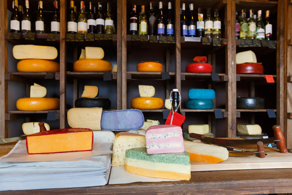 Grocery shop interior. Cheese wheels arranged on wooden shelves, coloured cheese and wrapping paper on counter and wine assortment, background